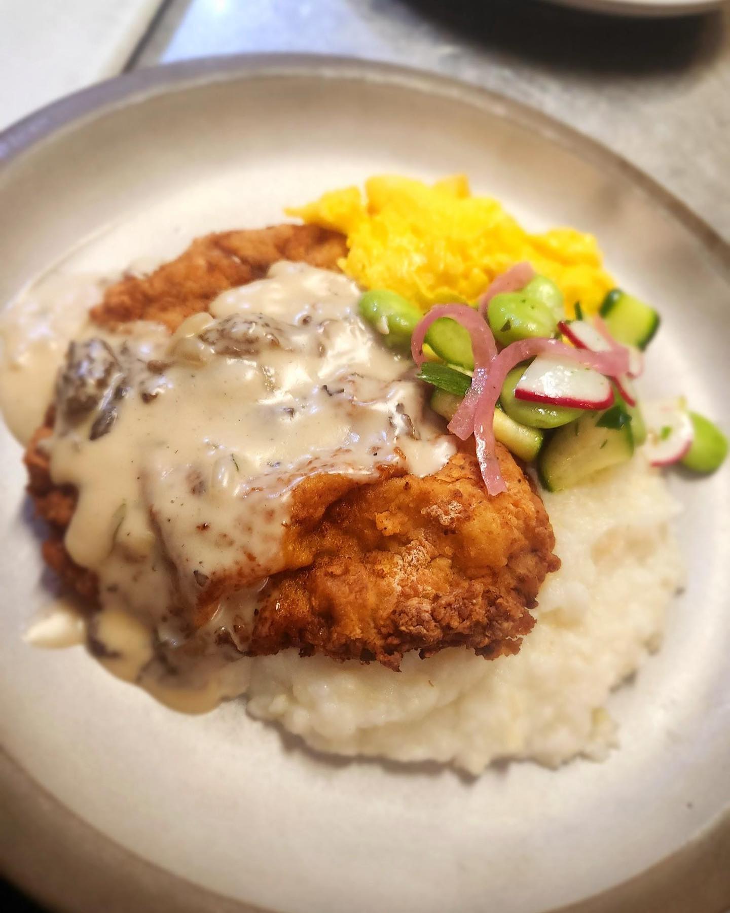 Crispy fried chicken topped with creamy gravy over cheese grits, with a cucumber, radish, and edamame salad on the side.